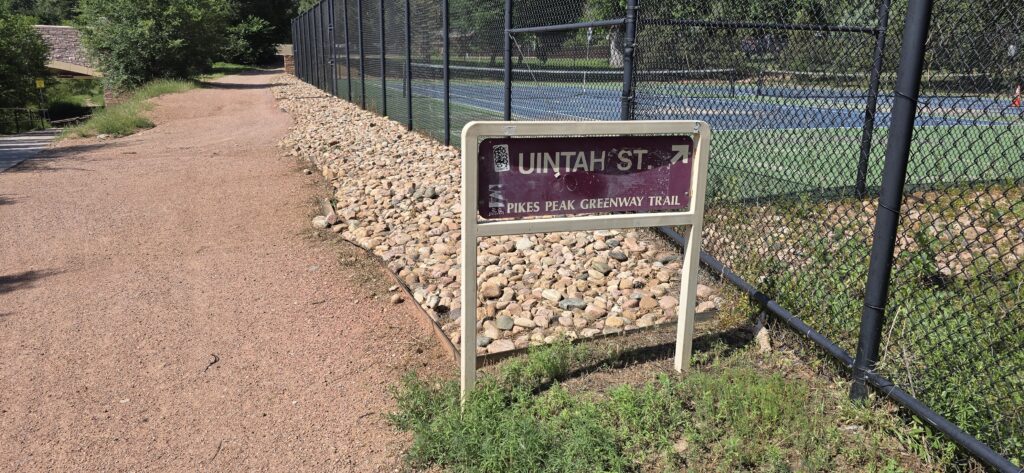 Maroon and tan sign for Pikes Peak Greenway along gravel path passing by a tennis court. In the vicinity of Uintah Street.