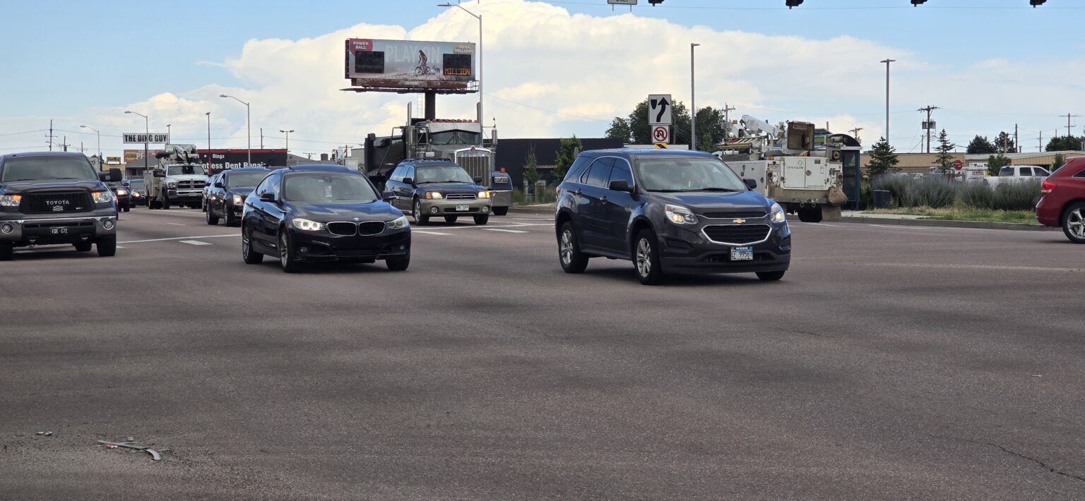 Traffic passing through intersection at Platte Ave and Circle Drive. A dump truck in the background behind vehicles.